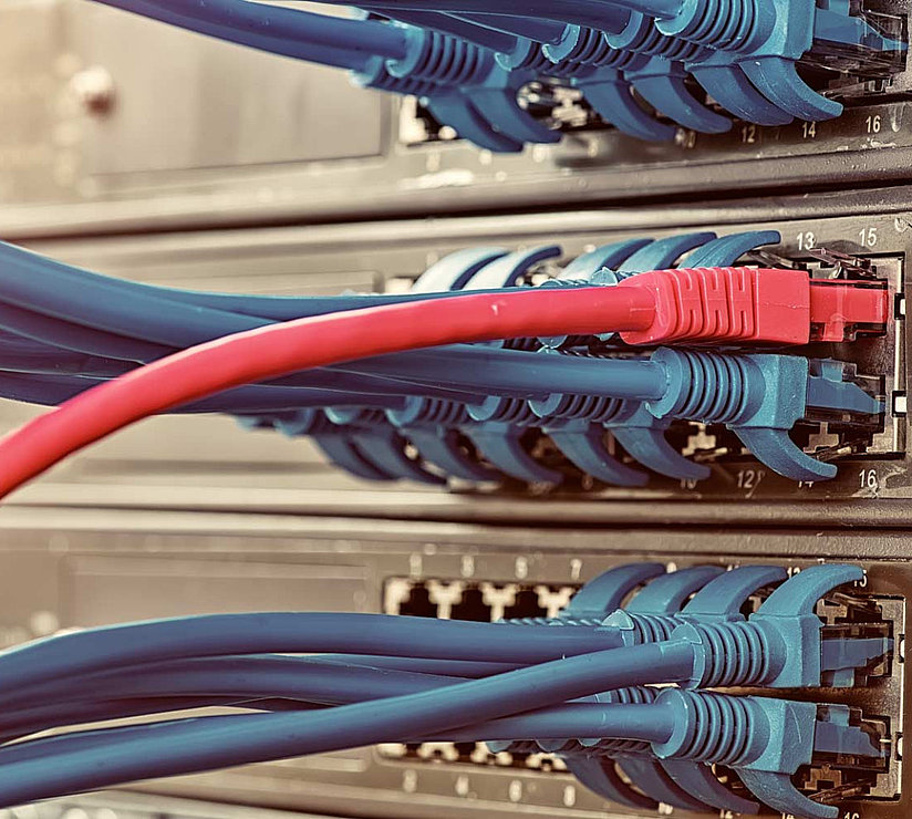 Nahaufnahme von Netzwerk-Switches mit mehreren blauen Ethernet-Kabeln und einem roten Kabel, das an die Ports angeschlossen ist, um die Computernetzwerkinfrastruktur und Datenverbindungen in einem Serverraum zu verdeutlichen.